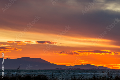 Sun has set down to the West with curly clouds near the fishermen village of Porto Lagos, Northern Greece. Mountain range and sea water in darkness. Blurred bushes in foreground