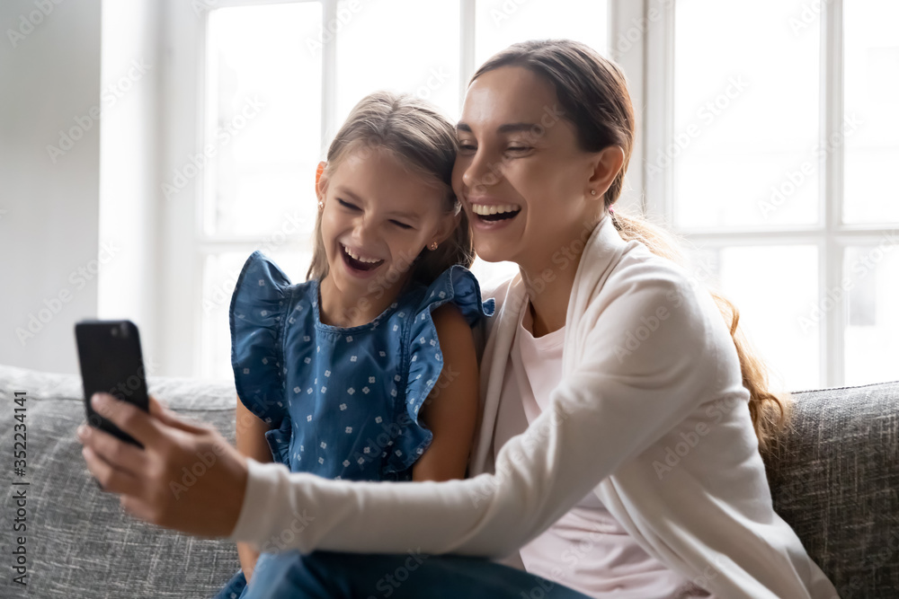 Overjoyed young Caucasian mom and small daughter sit on sofa at home laugh watching funny video on smartphone together, happy mother and little girl child have fun using cellphone relaxing on couch Overjoyed young Caucasian mom and small daughter sit on sofa at home laugh watching funny video on smartphone together, happy mother and little girl child have fun using cellphone relaxing on couch