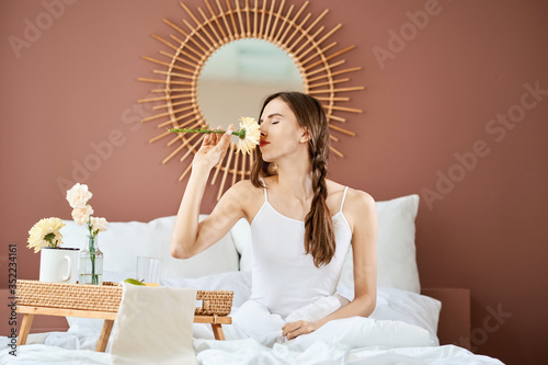 Fotografija Dreamy girl sitting on the bed and sniffing a flower