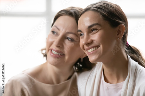 Close up of smiling millennial girl child and mature 60s mom look in distance dreaming of bright future together, happy middle-aged mother and grownup adult daughter think or visualize at home