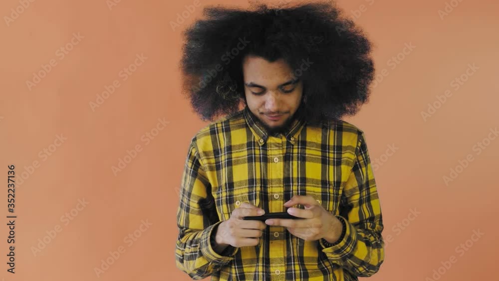Afro-American Man Playing Game on Smartphone on Orange background. Concept of emotions Afro-American Man Playing Game on Smartphone on Orange background. Concept of emotions