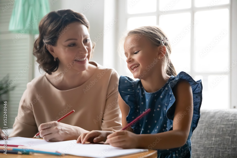 Caring middle-aged 60s grandmother sit at desk preparing homework handwriting with little granddaughter, loving senior granny study with small grandchild drawing painting together at home Caring middle-aged 60s grandmother sit at desk preparing homework handwriting with little granddaughter, loving senior granny study with small grandchild drawing painting together at home