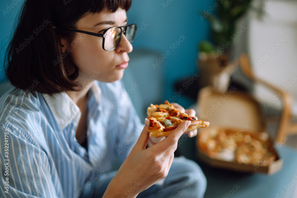 Young  girl eating a piece of pizza from the box, sitting at sofa at home.  Fast food. 