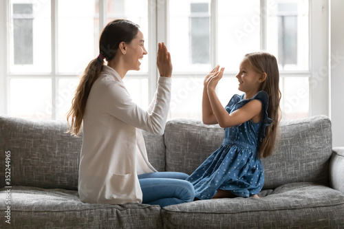 Overjoyed young mom and small daughter sit on sofa in living room play pat a cake hand clapping game together, happy mother and little preschooler girl child have fun enjoy family weekend at home