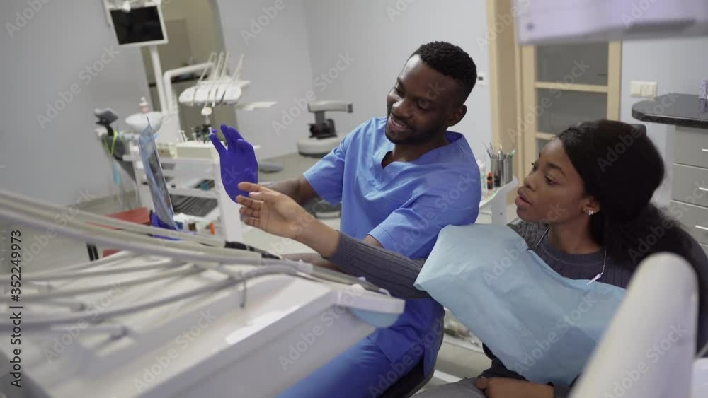 Dentisry concept, healthcare, oral and teeth care. Young handsome african man dentist discusses the strategy of treatment with the patient african charming young lady, showing xray image of jaws Dentisry concept, healthcare, oral and teeth care. Young handsome african man dentist discusses the strategy of treatment with the patient african charming young lady, showing xray image of jaws
