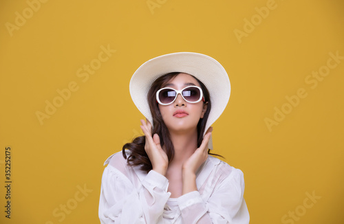 Portrait Asian beautiful happy young woman with sunglasses and hat smiling cheerful in summer and looking at camera isolated on yellow studio background.