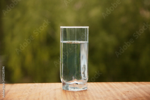 Pure cold water in a glass cup stands on a wooden table in the street against the background of green trees. Life style.