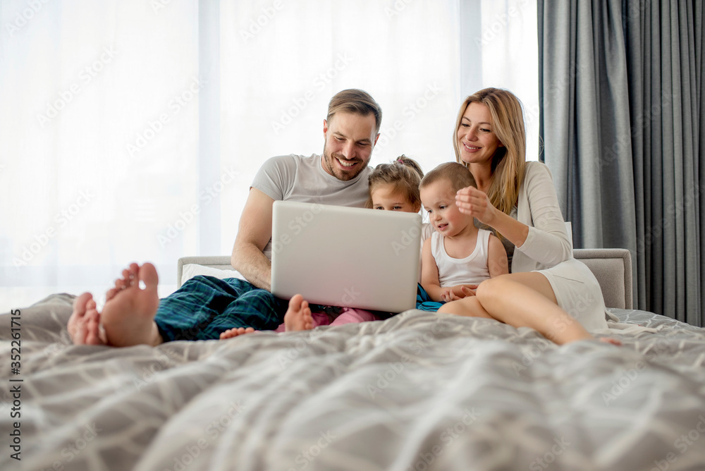 Happy family, parents and children, relaxing in bed together while watching funny videos and cartoons on laptop Happy family, parents and children, relaxing in bed together while watching funny videos and cartoons on laptop