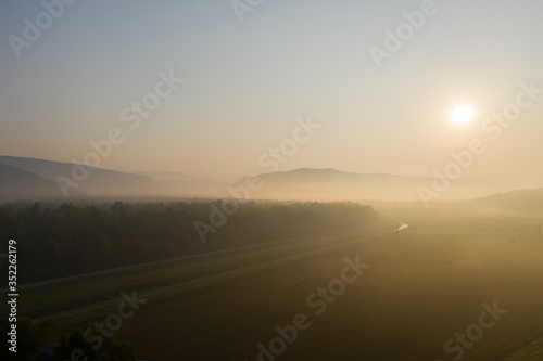 Rising sun. Mountain road along the edge of the forest in the thick morning fog. The view from the top. Shooting from a drone.