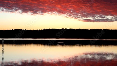Mirror image of pink light sky with bright burning crimson clouds in the lake at dawn.The illuminated black forest on the horizon is copied in the calm water.A glossy picture in the twilight.Russia