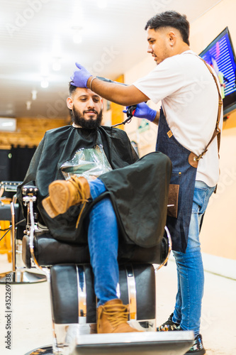Young Latin Barber At Work In Stylish Barbershop. Cool, bearded man enjoys the moment.