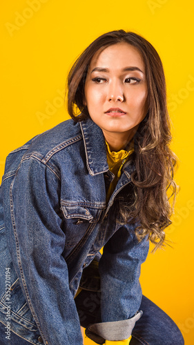 a girl with brown wavy hair sits on a yellow background looking to the side, in a blue jeans, Asian appearance
