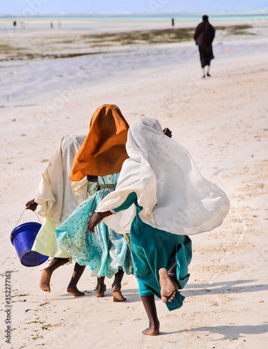 3 Muslim youngsters with colorful clothes and head covers running on the beach in Zanzibar, with a woman in the background.