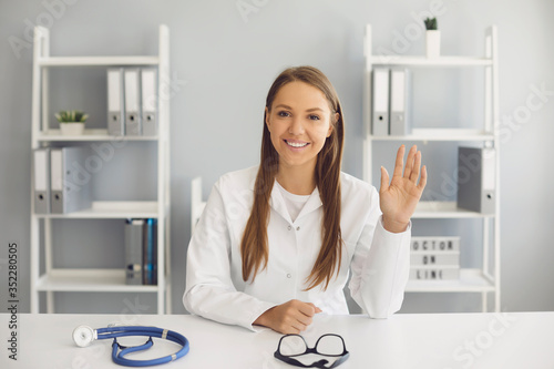 A female doctor looking at the camera has a video chat sitting gretting in a clinic office. Remote medical consultation. photo