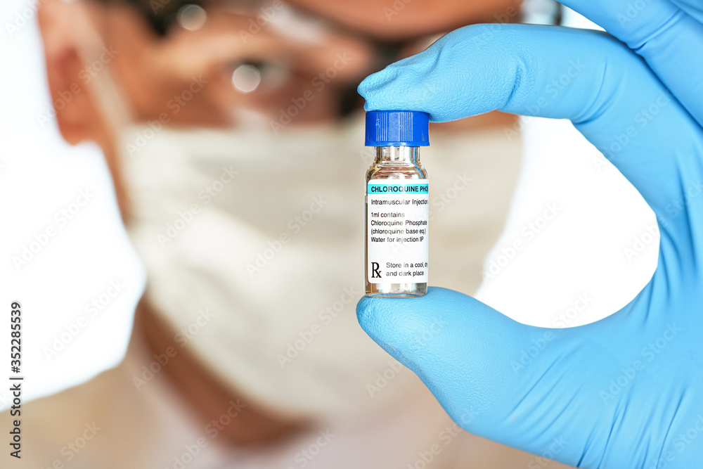 Hand in blue medical gloves holding small vial with Chloroquine Phosphate (generic name, own design label), blurred face in cotton mask and goggles background. Potential covid 19 coronavirus cure