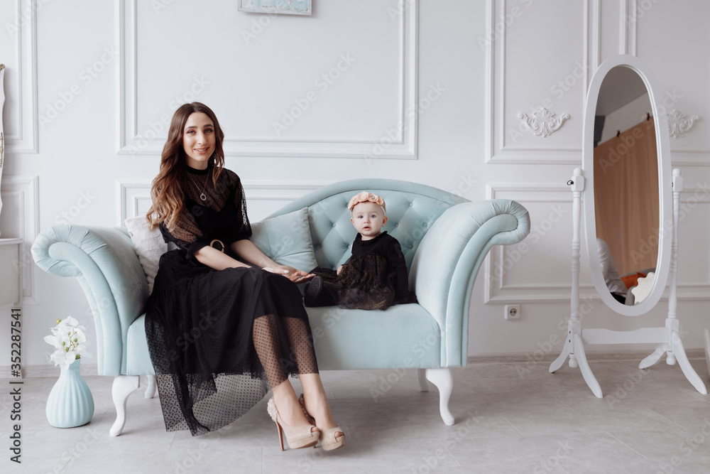 Happy young mom on stylish couch relax with little daughter in black dresses and posing, smiling mother and girl child have fun at studio. family look Happy young mom on stylish couch relax with little daughter in black dresses and posing, smiling mother and girl child have fun at studio. family look