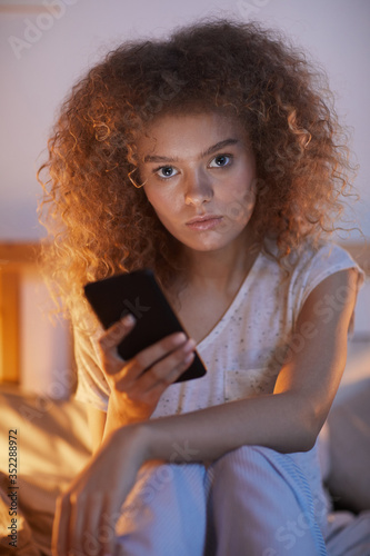 Vertical portrait of young curly-haired woman using smartphone while sitting on bed at night and looking at camera