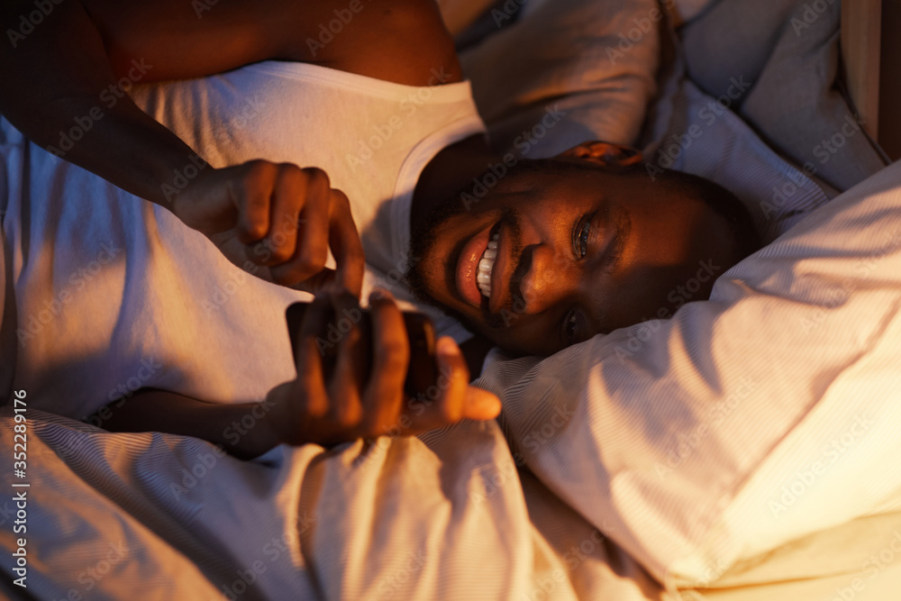 Obraz premium High angle portrait of smiling African-American man using smartphone in bed at night while texting girlfriend, copy space