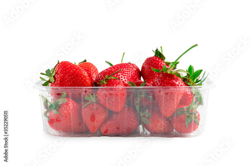 Heap of vivid saturated ripe red strawberries laying in big plastic transparent container and one big berry laying near the box isolated on clear white background