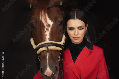 Smiling jockey woman standing with brown horse in stable, black background