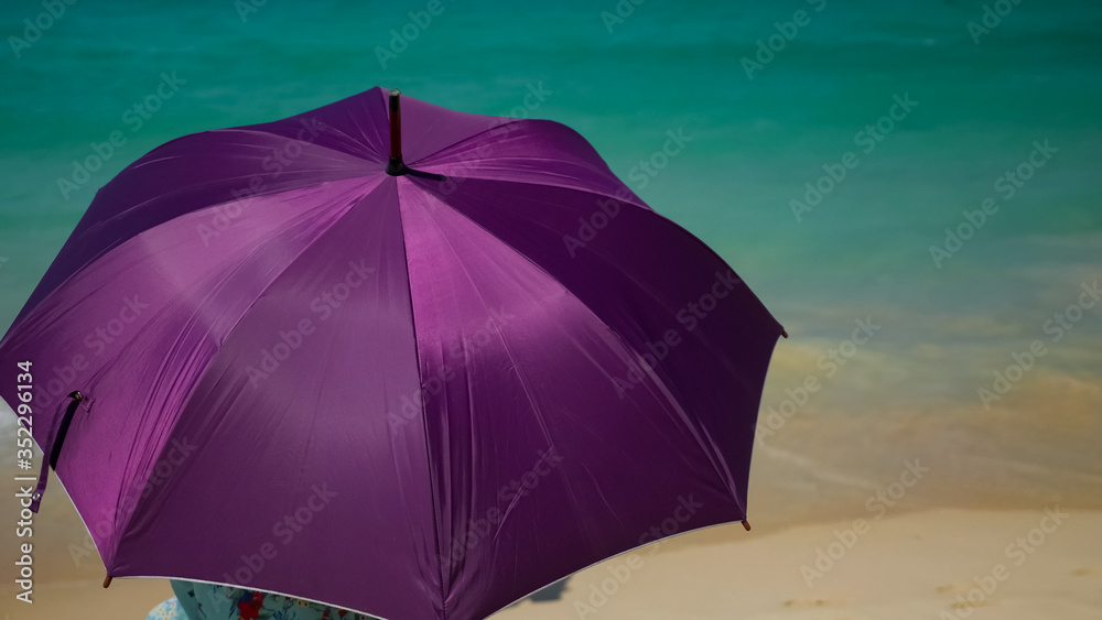 woman in dress with flower pattern sits on yellow beach holding purple umbrella hiding from sun against ocean backside view