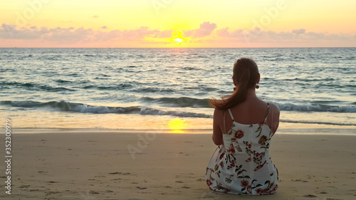 young woman in stylish summer dress sits on beach sand watching pictorial ocean at sunset time backside view