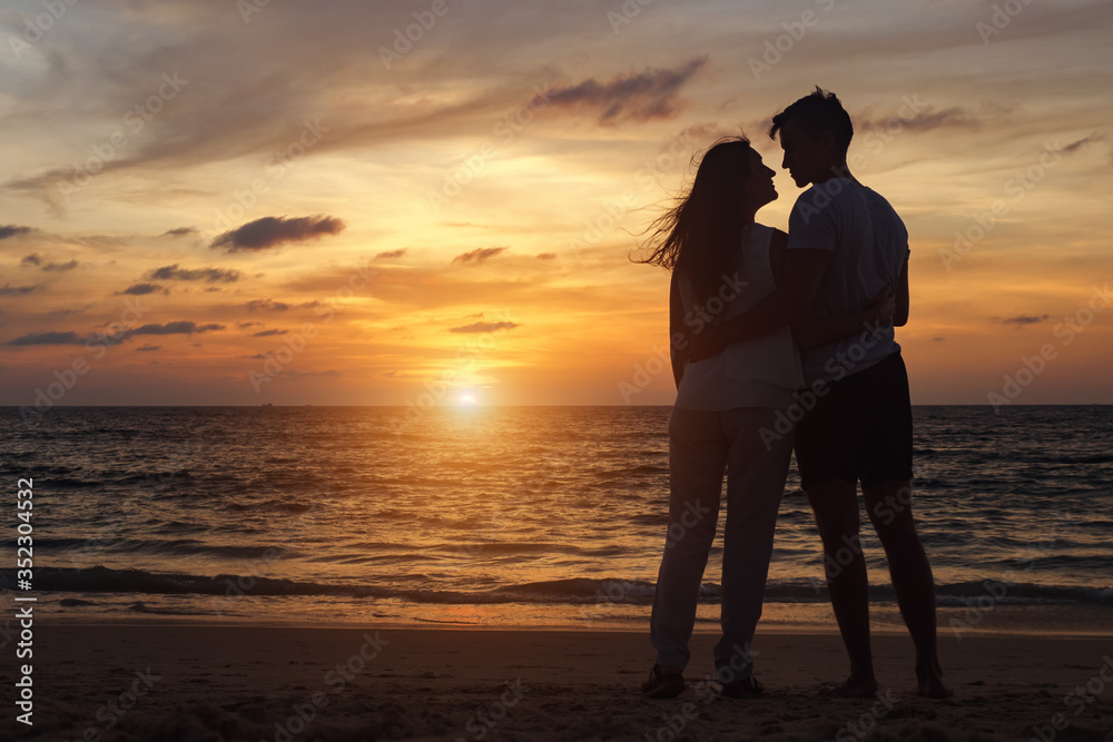 silhouette of young couple standing on beach looks at each other at orange sunset backside view copy space