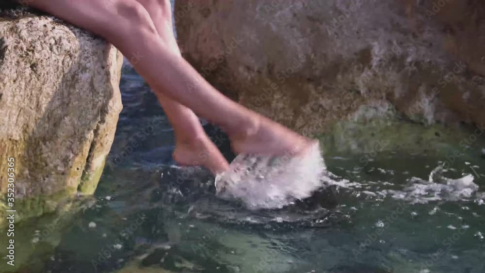 Tanned pretty legs splashing with sea water. A young woman sits on a large stone, her legs dangling in the transparent blue water, the sun is shining. Tanned pretty legs splashing with sea water. A young woman sits on a large stone, her legs dangling in the transparent blue water, the sun is shining.