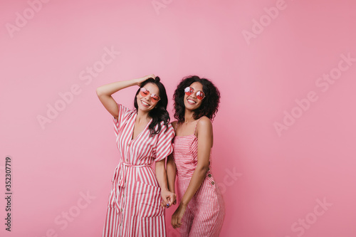 Relaxed caucasian girl playing with dark hair while posing with cute african lady. Indoor shot of stylish female models isolated on pink background