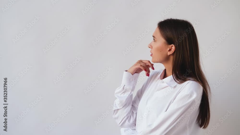 Beautiful young woman in a white shirt looks away and smiles, she laughs. Girl with brown hair and tanned skin playfully posing against a white wall. Beautiful young woman in a white shirt looks away and smiles, she laughs. Girl with brown hair and tanned skin playfully posing against a white wall.