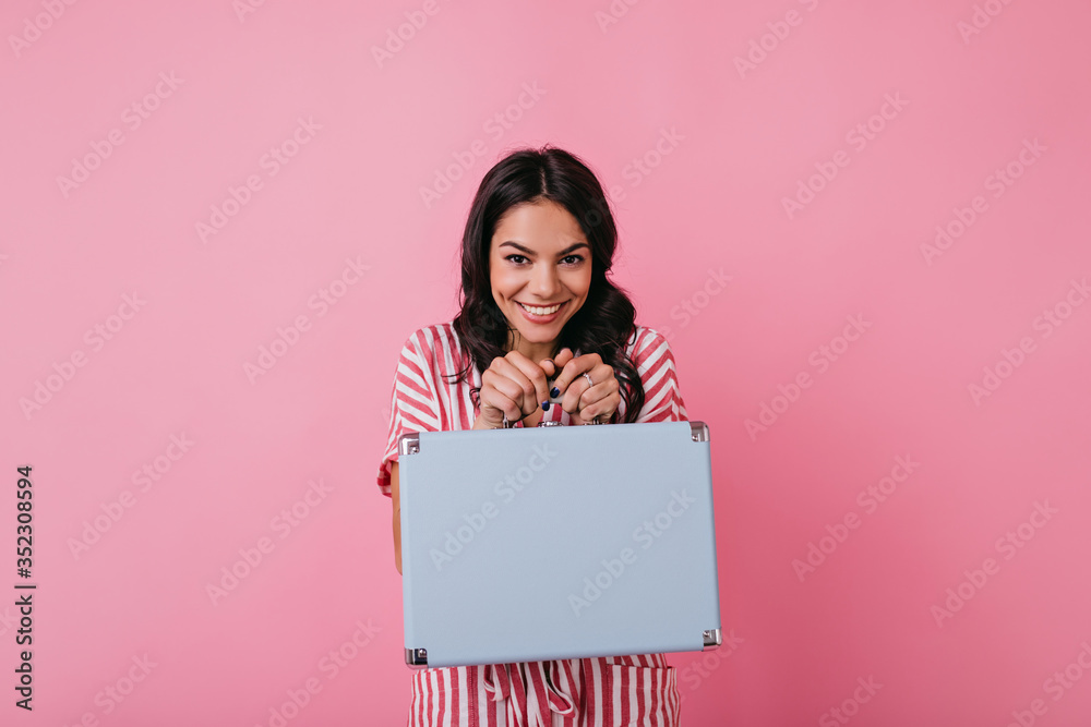 Portrait of charming girl in striped outfit holding light blue suitcase. Brunette with friendly smile poses on pink background Portrait of charming girl in striped outfit holding light blue suitcase. Brunette with friendly smile poses on pink background
