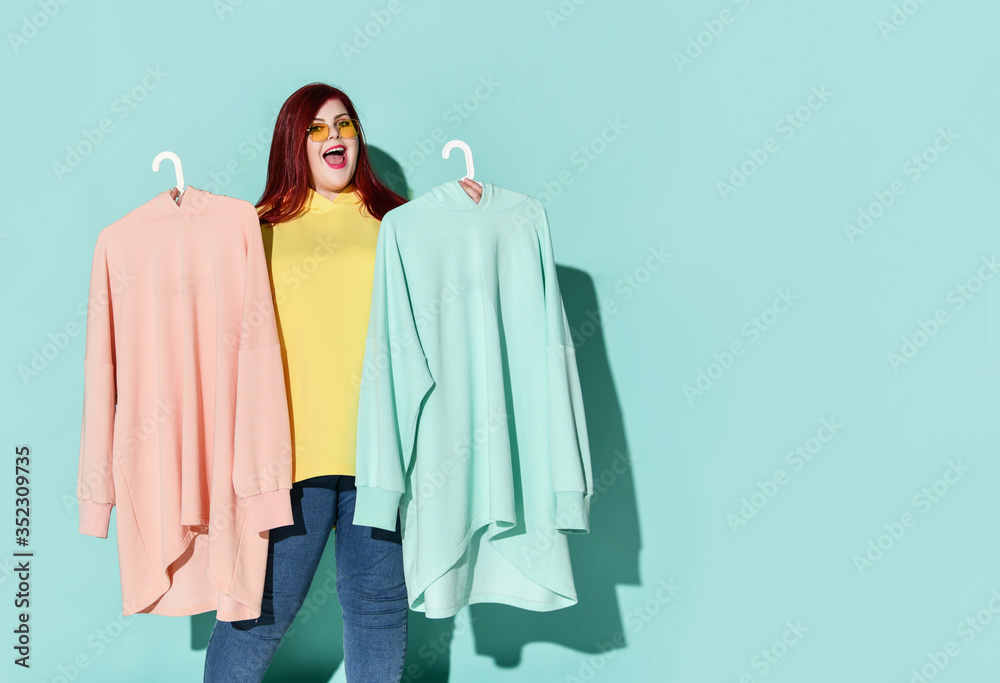 Young smiling red haired overweight woman in casual clothing standing and choosing between blue and pink hoodie Young smiling red haired overweight woman in casual clothing standing and choosing between blue and pink hoodie