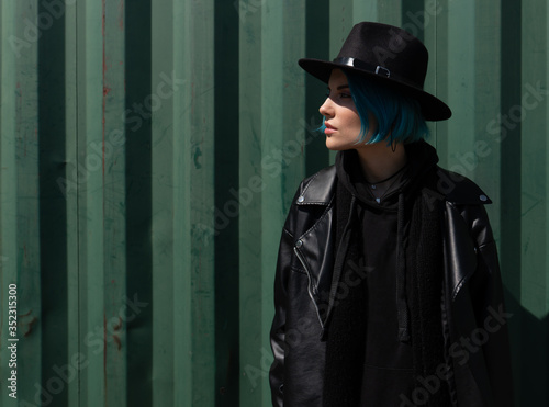 Girl with blue hair in a black hat and leather jacket poses near a green container. Copy space © Artem