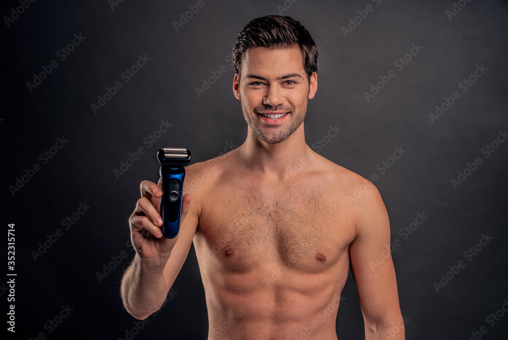 Handsome young bearded man isolated. Portrait of topless muscular man is standing on gray background with electric Shaver in hand. Men care concept.