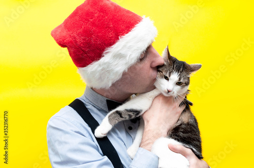 handsome man in red santa hat holding and kissing cute tabby cat in front of yellow background
