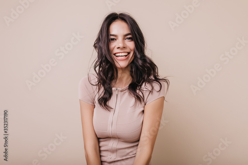 Portrait of cute dark-haired girl with snow-white smile. Brown-eyed lady in beige top looking at camera
