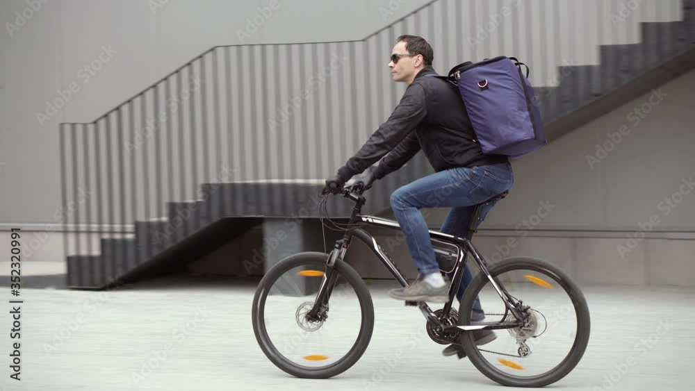 Courier delivery service. A man in glasses and a windbreaker rides a bicycle with a backpack along a city street against the background of the building. Online food and goods orders Courier delivery service. A man in glasses and a windbreaker rides a bicycle with a backpack along a city street against the background of the building. Online food and goods orders