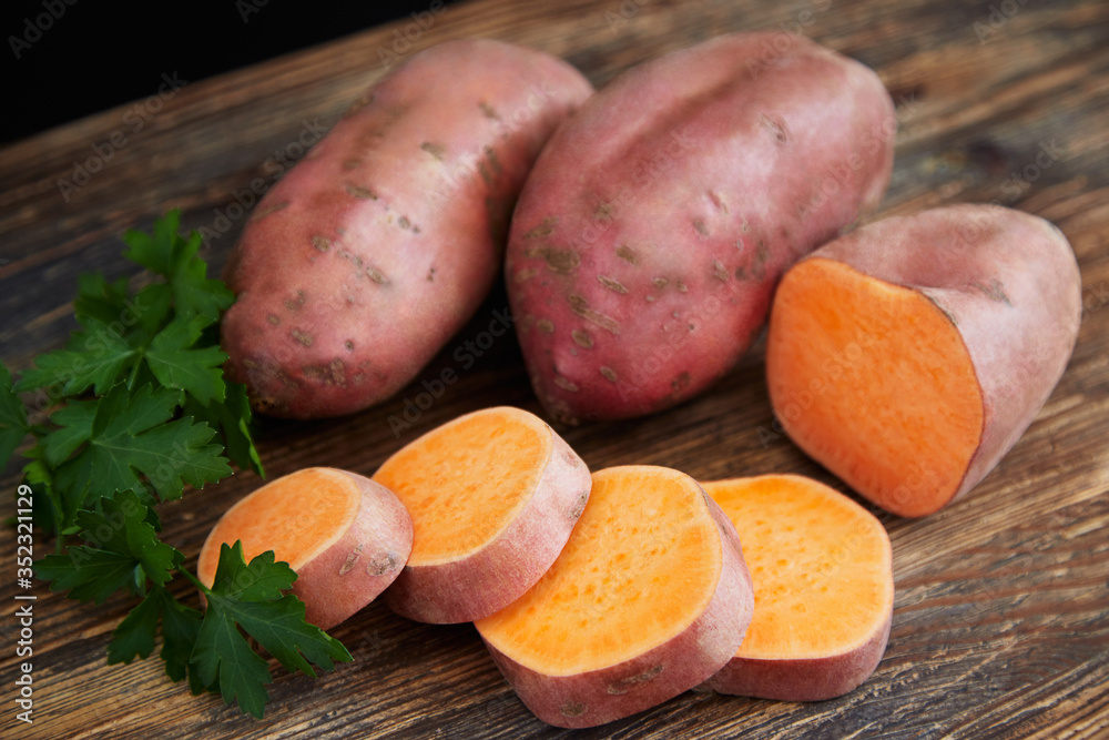 Raw sweet potatoes (batatas) on a rustic wooden table decorated with parsley. One batata in the front is half sliced. Raw sweet potatoes (batatas) on a rustic wooden table decorated with parsley. One batata in the front is half sliced.