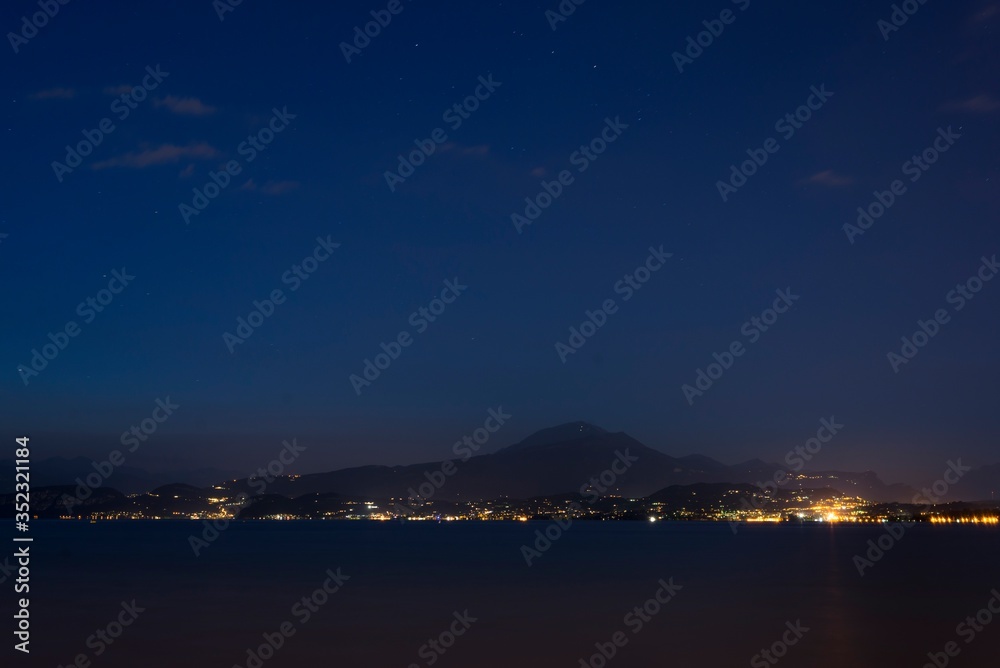 Night shot from a coast of Peschiera del Garda to the Garda lake and mountains with lights all over the other coast. Lights of Garda town in the night from other coast.