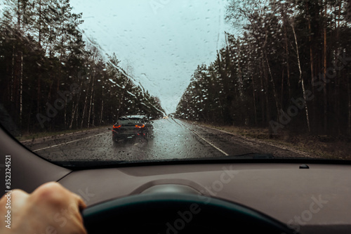 View through windshield of car on drops on glass, wet road and rainy weather. Highway car traffic in bad weather, downpour. Dangerous overtaking. Dashboard inside and driver hand with steering wheel photo