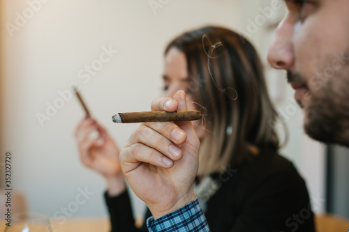 Side portrait of a white man with a beard smoking Cuban cigarettes. A woman in the background with a cigarette
