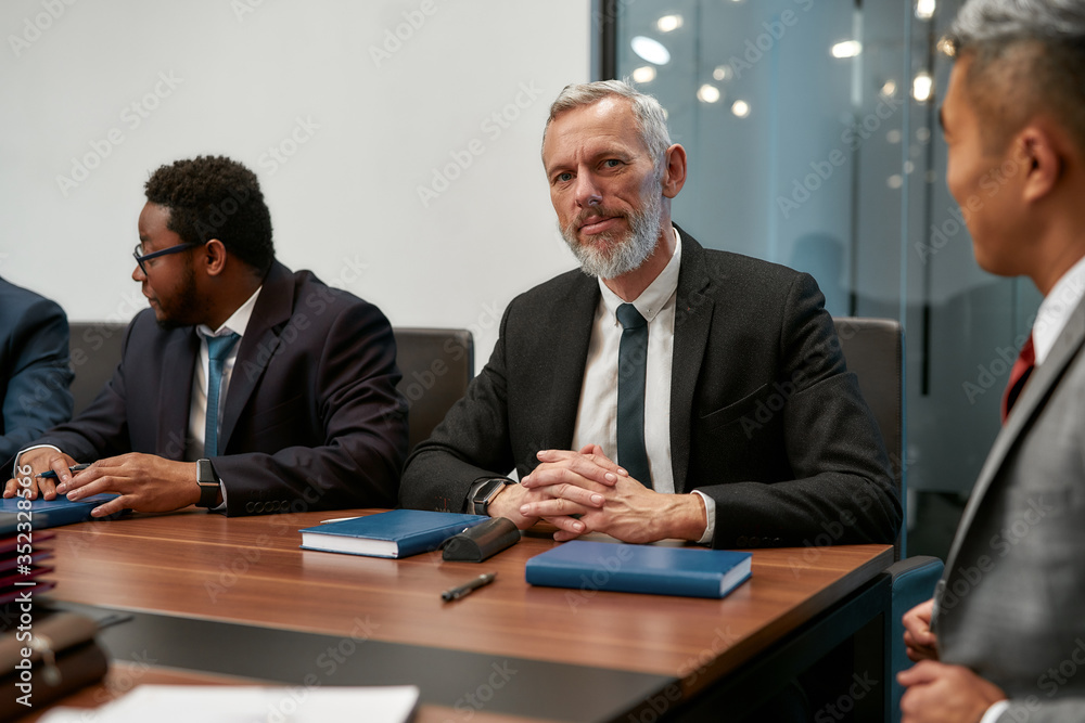 Busy working day. Portrait of bearded mature man in formal wear looking at camera while sitting at the office table with his co workers in the modern office Busy working day. Portrait of bearded mature man in formal wear looking at camera while sitting at the office table with his co workers in the modern office