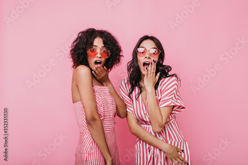 Shocked brunettes in bright summer dresses posing looking at camera. Portrait of attractive american models in pink studio