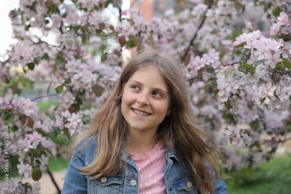 Portrait of a cute smiling young girl with blond hair in a blossoming spring garden. Girl dressed in denim jacket. Portrait of a cute smiling young girl with blond hair in a blossoming spring garden. Girl dressed in denim jacket.