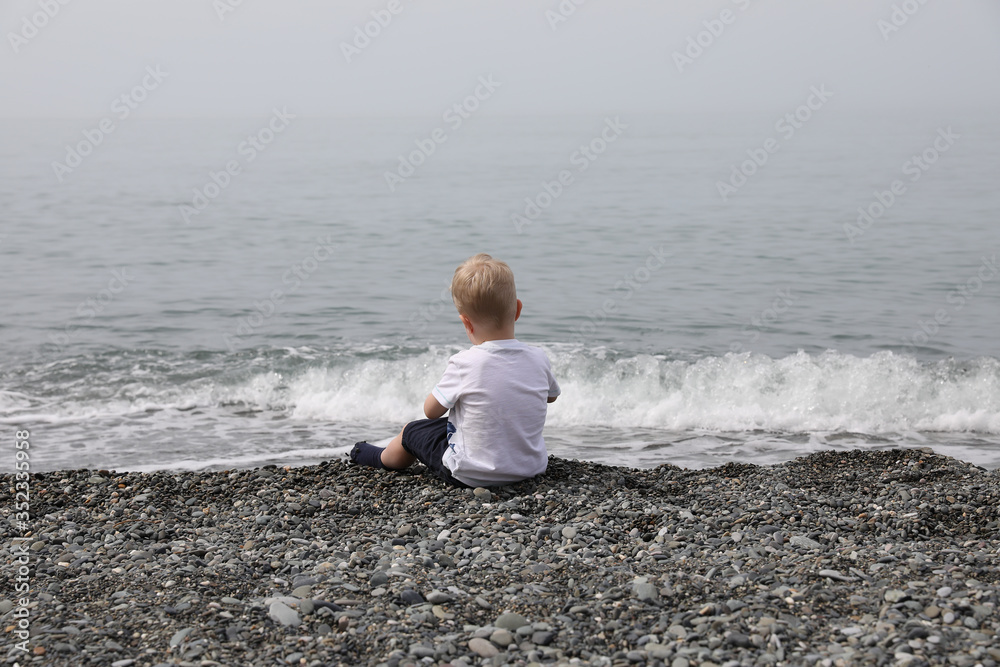 A little boy with blond hair sits on a pebble by the sea.