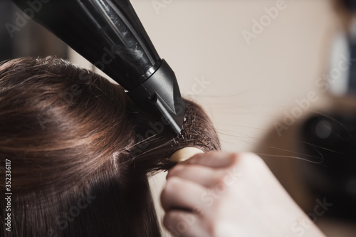 a professional hairdresser using round brush and blow dryer, styling long blond hair of his female client.