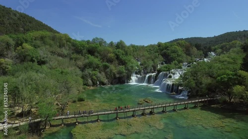 Aerial shot of tourists on footbridge over river near waterfall, drone descending over green water amidst plants and trees in forest against sky - Krka, Croatia photo