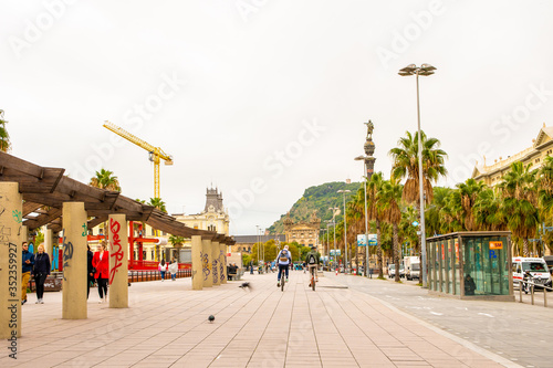 Barcelona, Spain. Wide sidewalk in La Rambla with men cycling, people walking and traffic on one way street beside. Columbus monument, Agencia Tributaria & a tower crane in distance