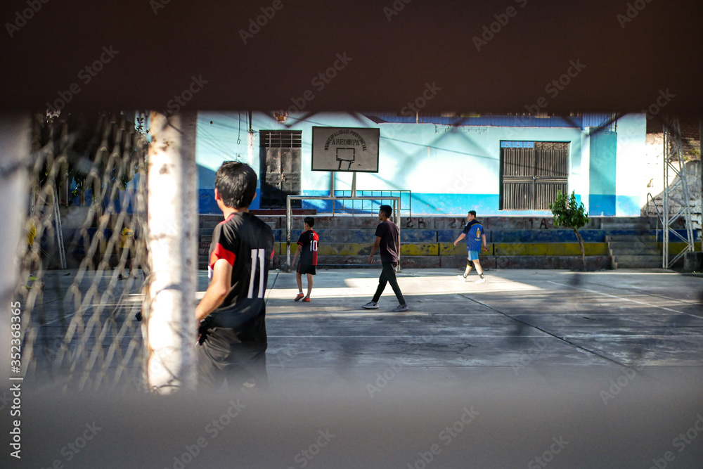 Fototapeta premium View on the group of young boys playing football at the concrete playground at the school campus behind the fence.