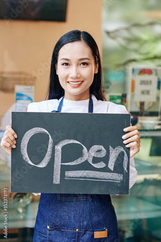 Positive young Asian woman in denin apron standing in cafe and showing open sign photo